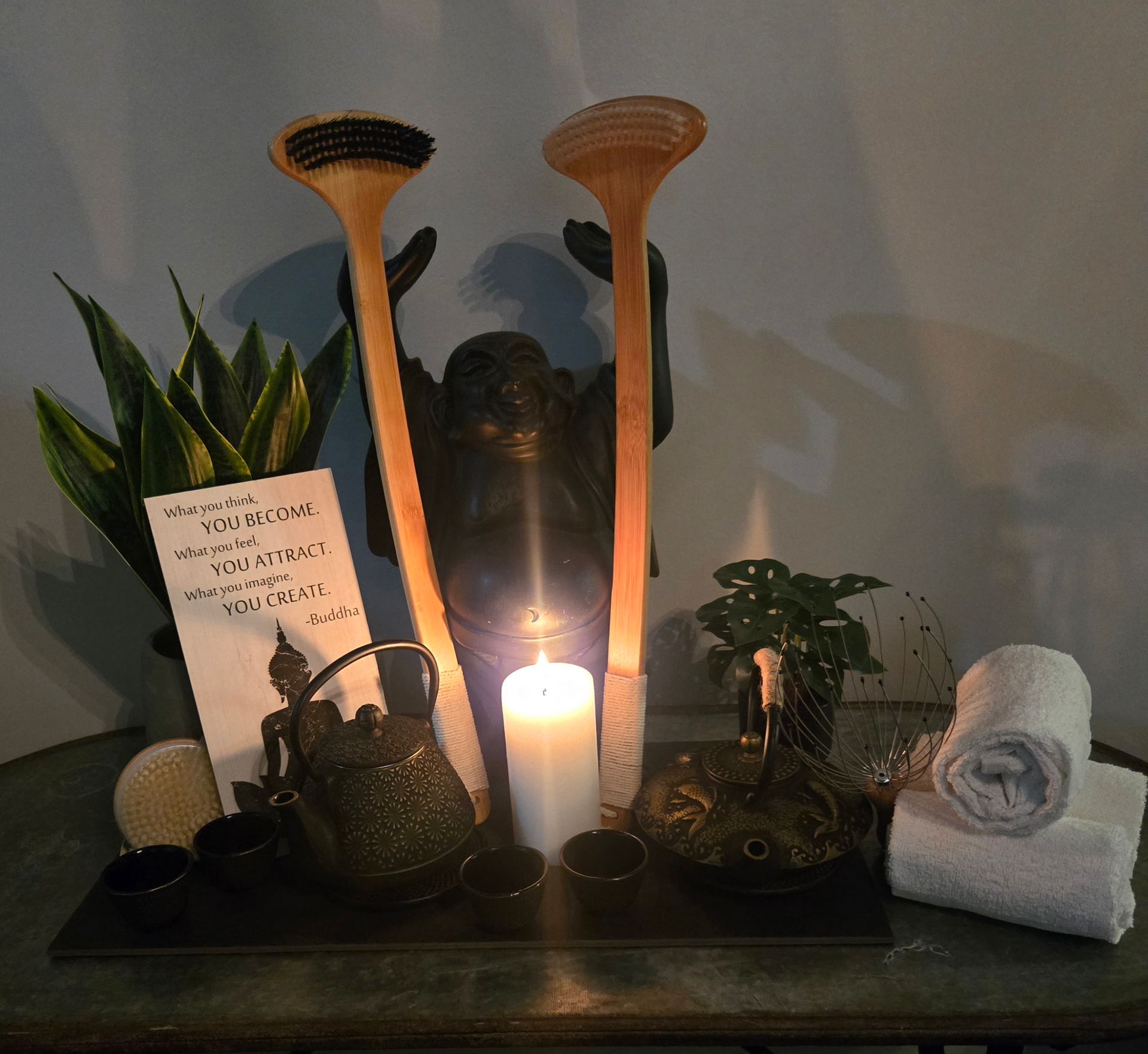 A spa scene: a lit candle, tea set, folded towels, plants, wooden massage tools, and a Buddha statue.