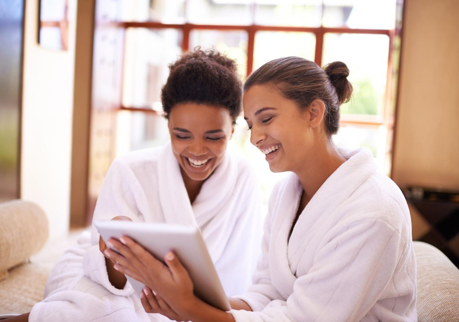 Two people wearing white bathrobes laugh while looking at a tablet together in a bright, indoor setting.