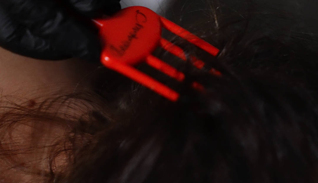 A gloved hand uses a red hair pick to separate dark, curly hair.