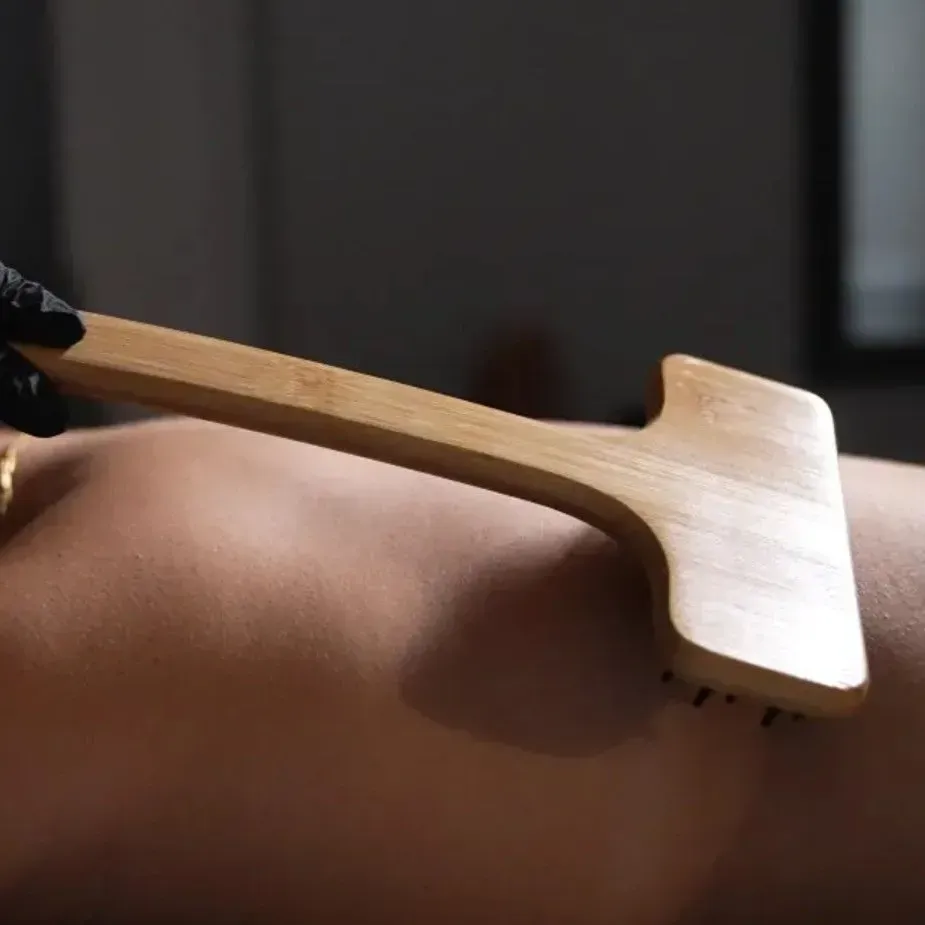 A gloved hand uses a wooden, T-shaped massage tool to rub a person's back.