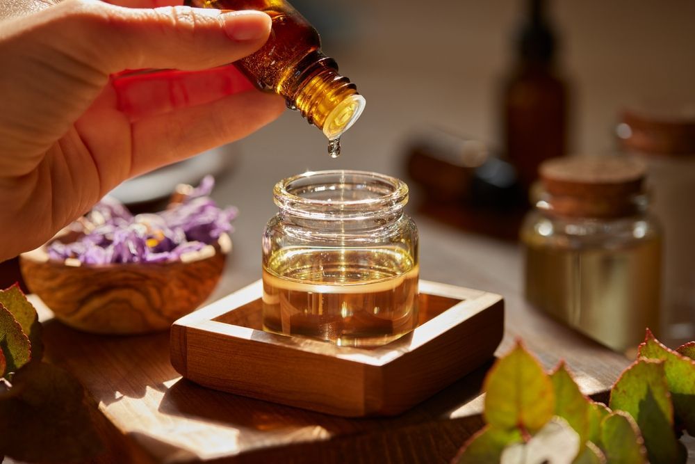 A hand adds a drop of golden essential oil from a small amber glass bottle into a clear jar on a wooden tray.