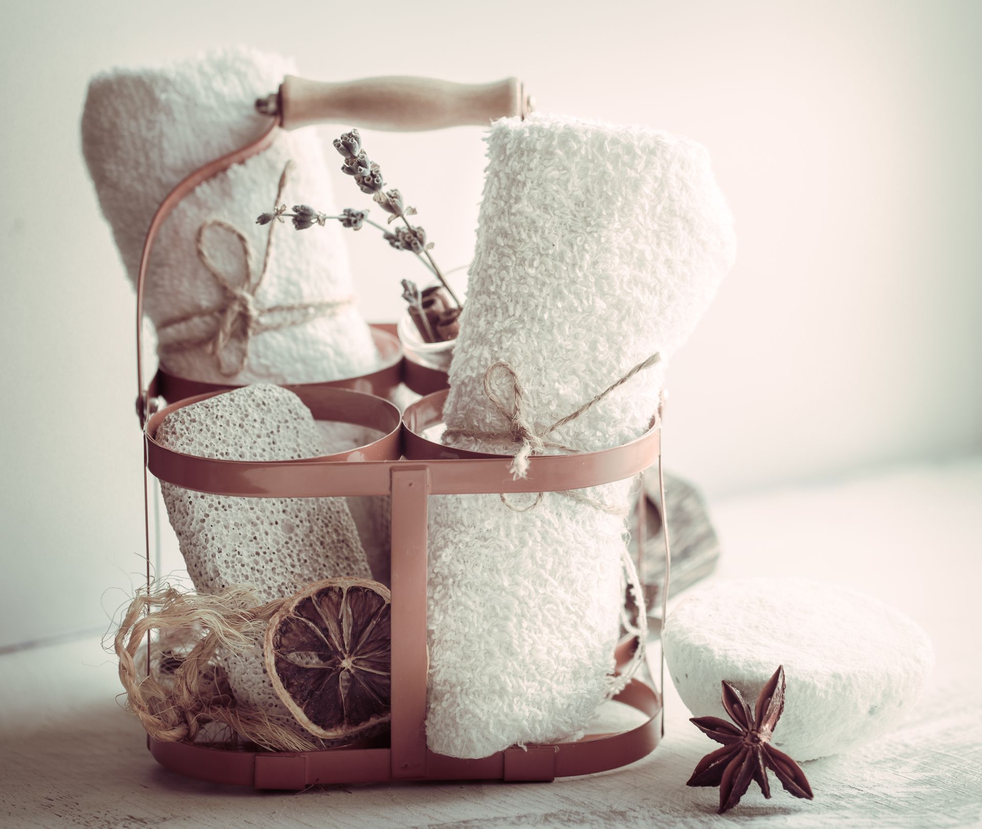 Basket with folded white towels, loofah, dried orange, and lavender sprig; spa setting.