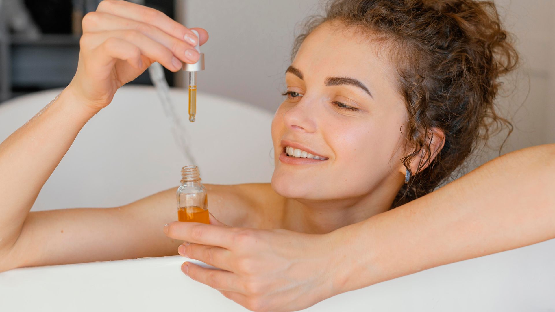 Woman in bathtub applying skincare serum with a dropper; smiles.