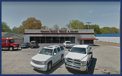 A body shop building with white walls and a large, paved parking lot featuring several cars and a red truck.