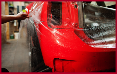 A person applying a clear protective paint film to the rear quarter panel of a red car in a garage.