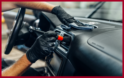 A person wearing black gloves uses a small brush and a cloth to clean the air vent of a car dashboard.