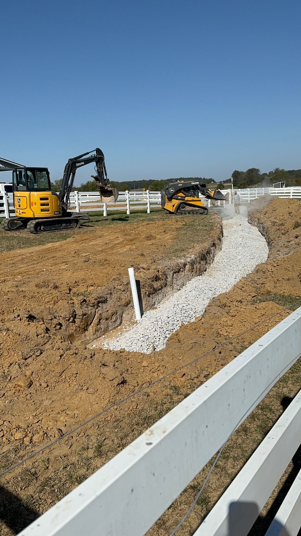 A yellow excavator is digging a trench in the dirt next to a white fence.
