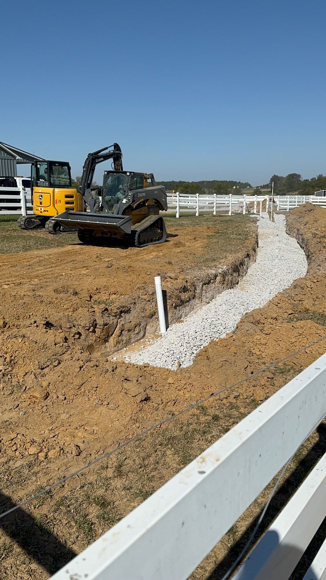 A bulldozer is digging a trench in the dirt next to a white fence.