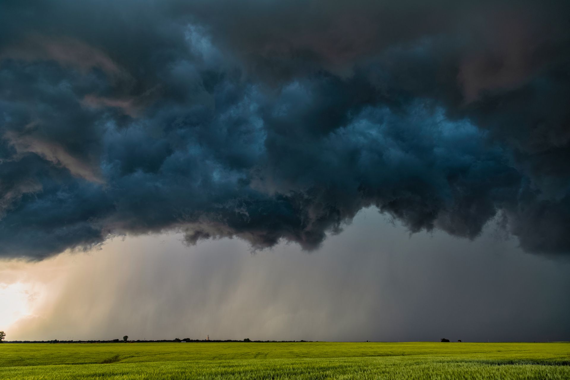 A large storm cloud is moving over a green field.