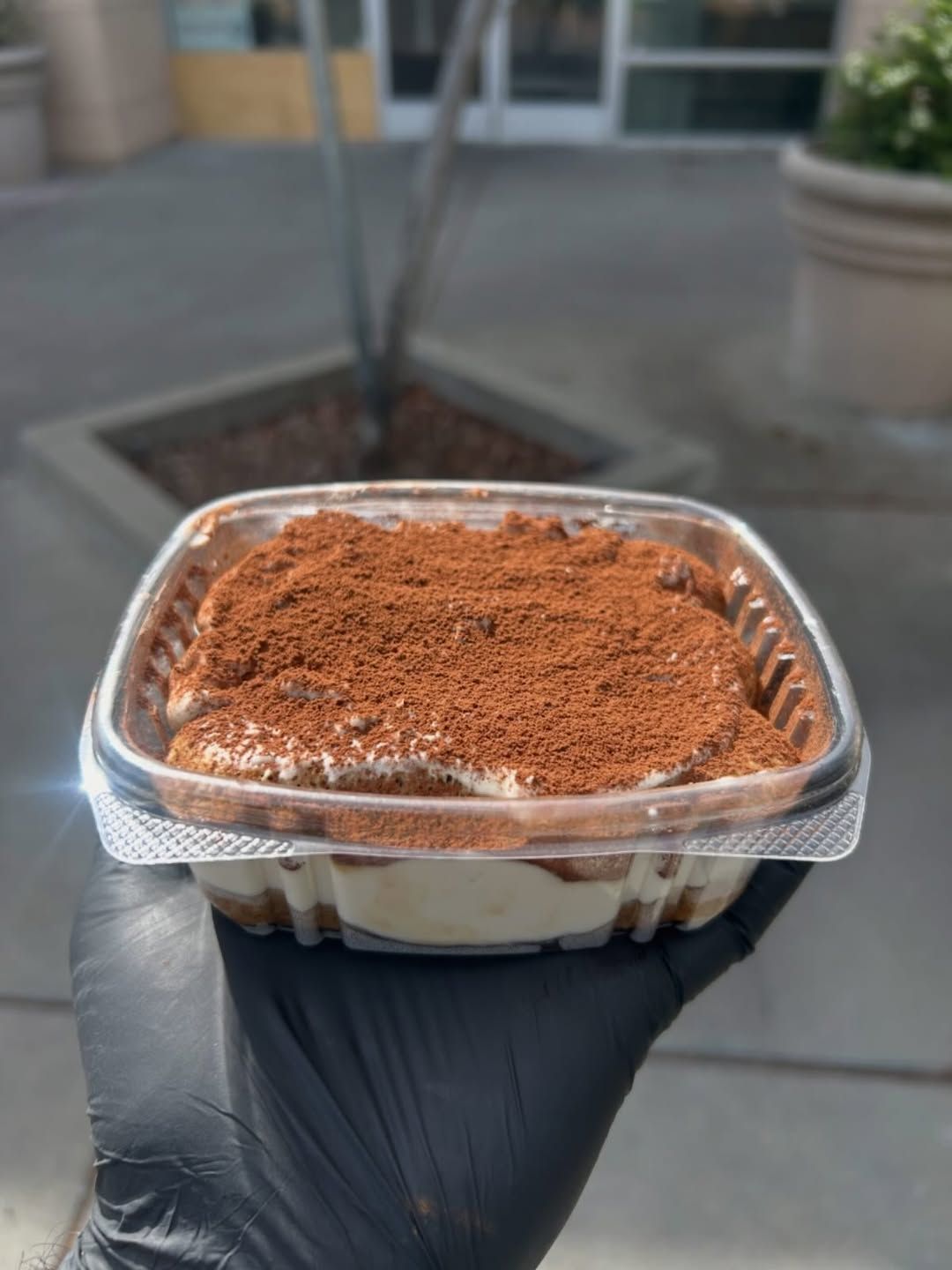 Hand holding a clear plastic dessert container topped with cocoa powder, outdoors on a sidewalk.