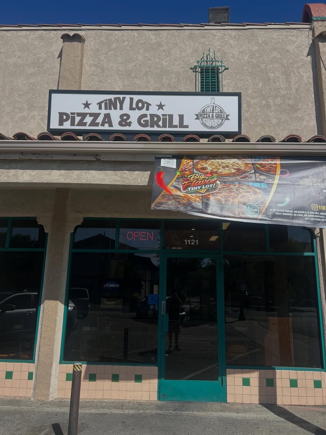 Storefront sign for Tiny Lot Pizza & Grill with teal doors and a colorful banner above the entrance