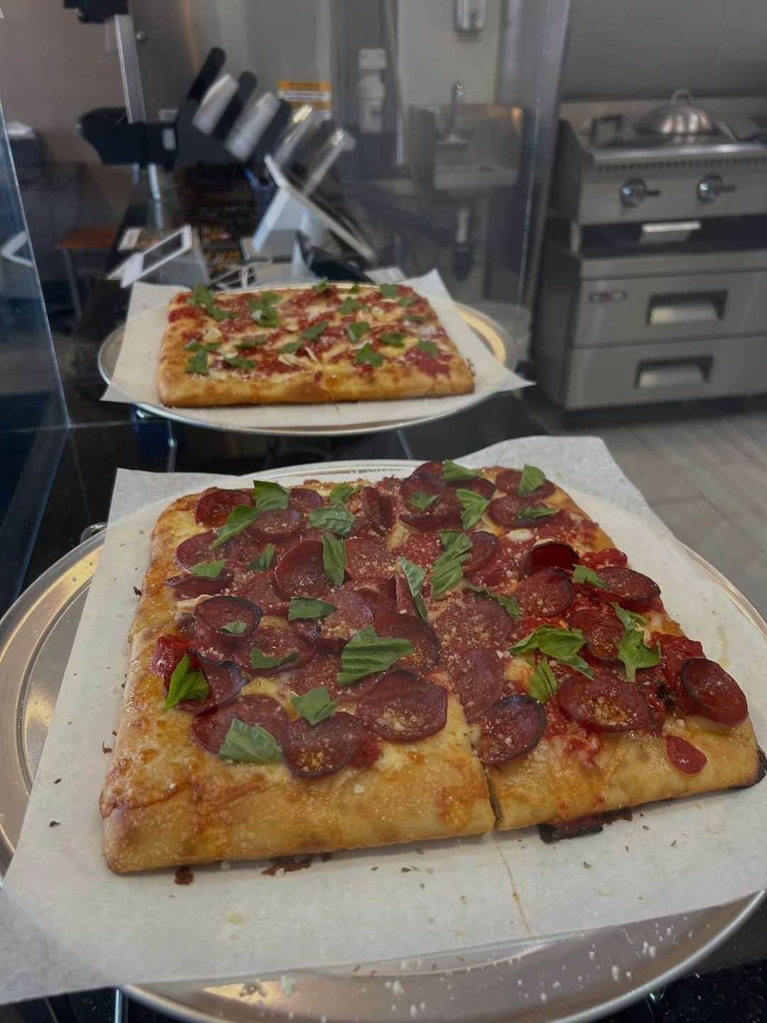 Two square pizzas on a kitchen counter, one pepperoni and basil in the foreground, one veggie in back