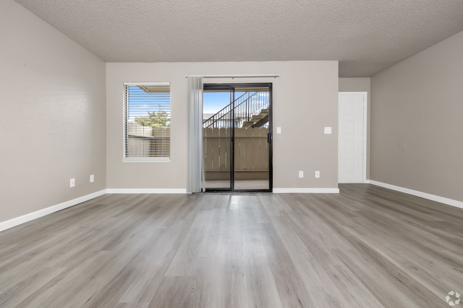 Empty living room with light grey walls, wood-look flooring, sliding glass door, and a window.