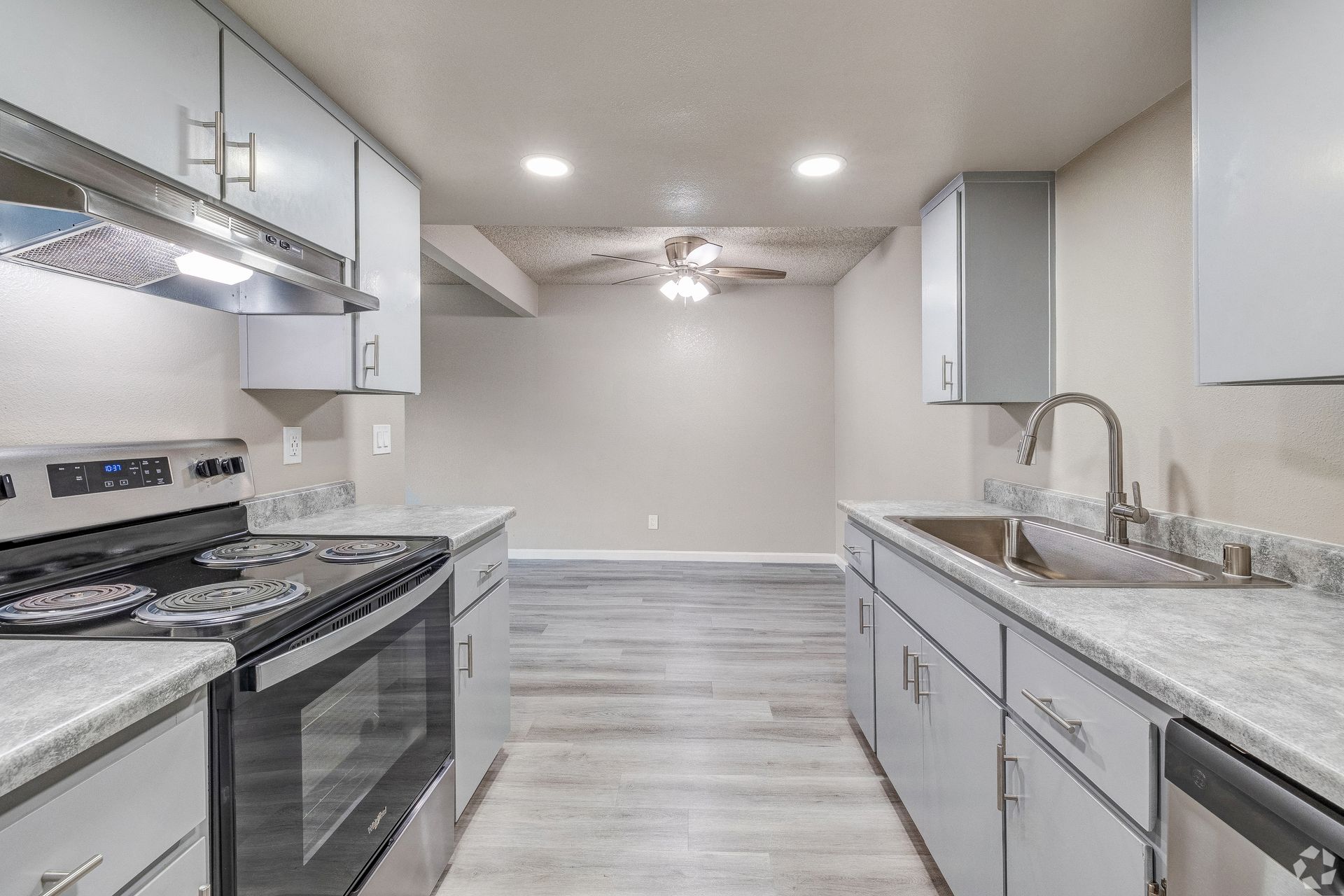 Kitchen with grey cabinets, stove, sink, and granite countertops. View into dining area with light floors.