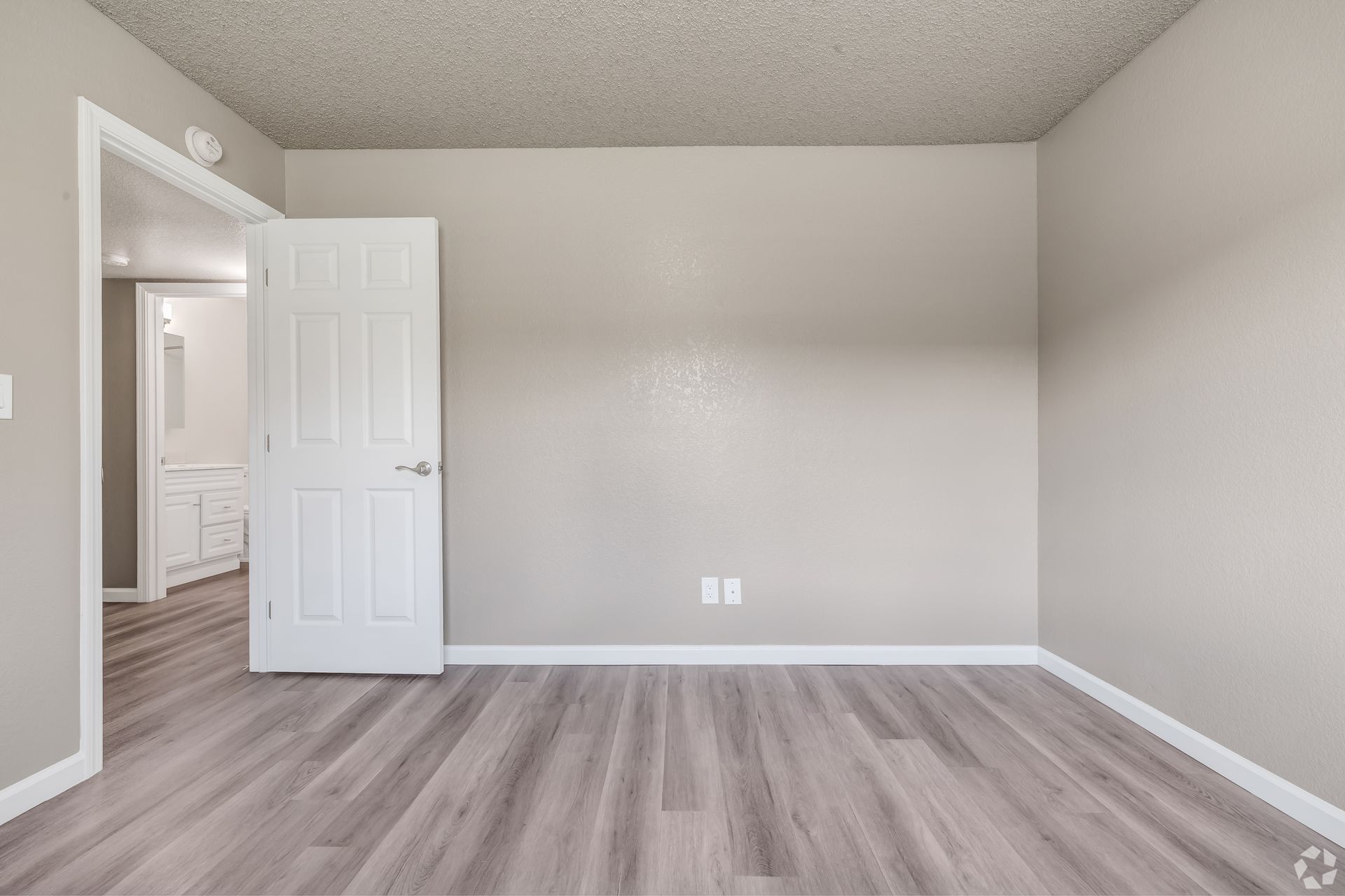 Empty bedroom with gray wood-look floors, beige walls, and a white door leading to a bathroom.