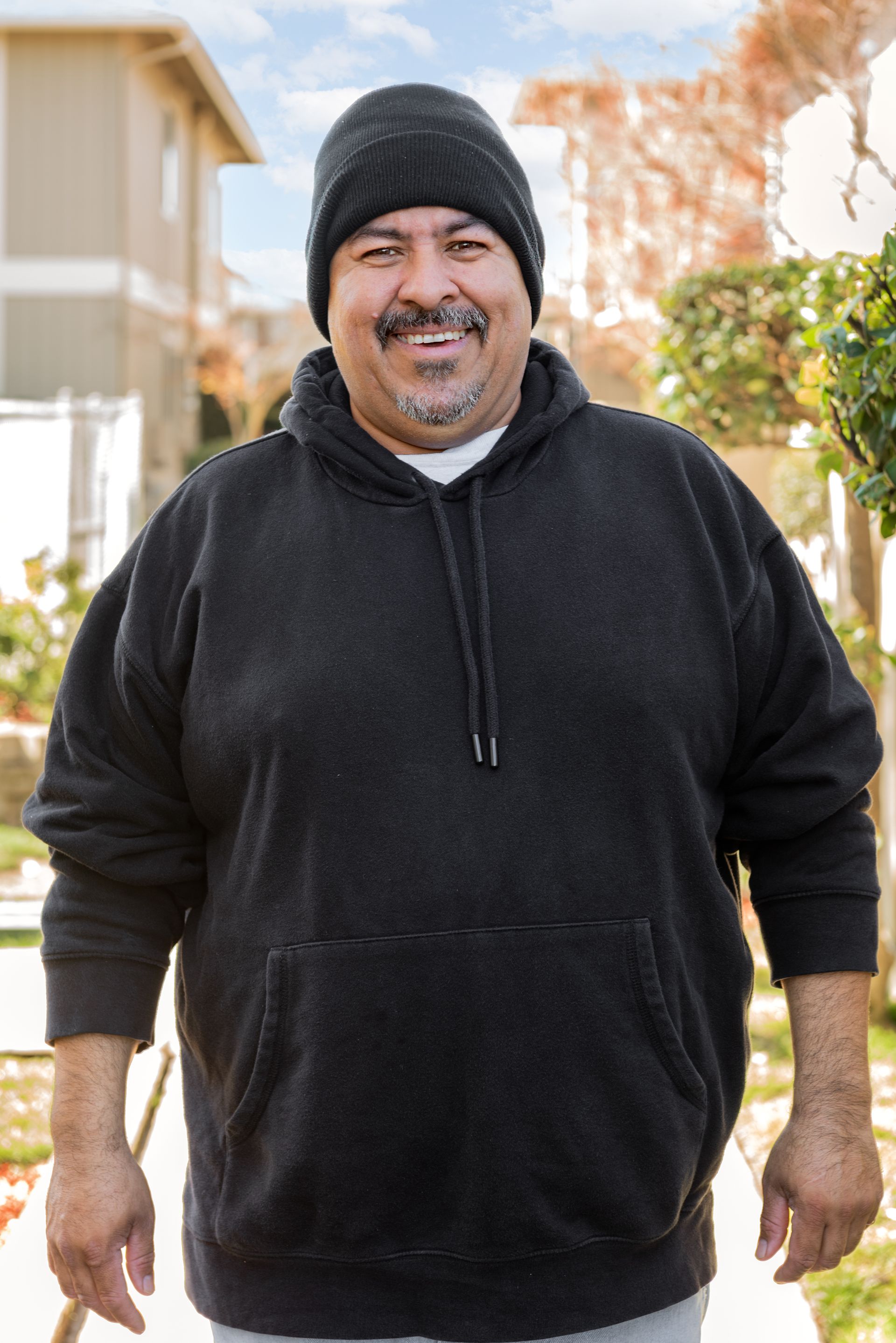Man in black hoodie and beanie smiles outdoors.