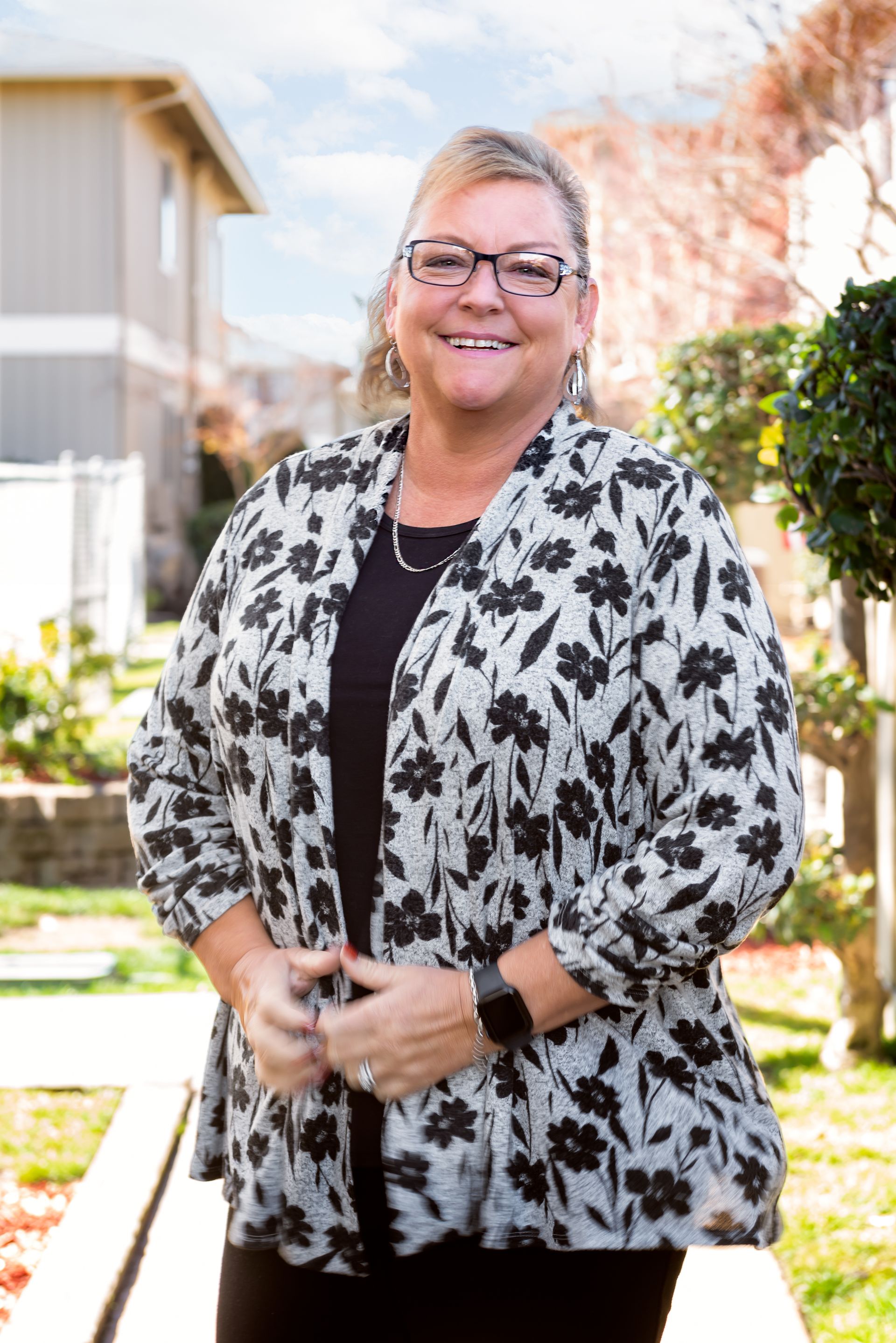 Woman smiling, wearing glasses, floral jacket, standing outdoors.