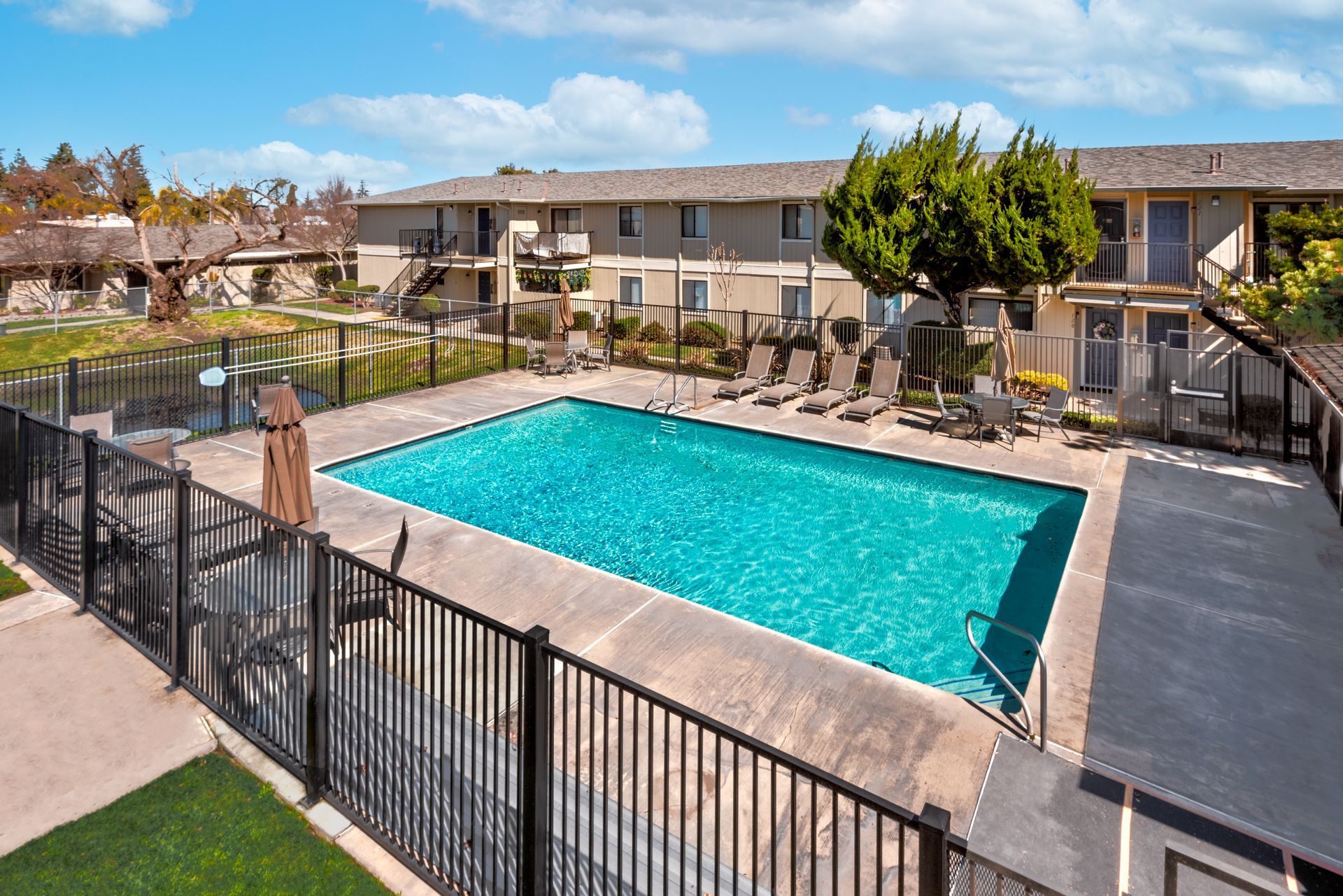Pool surrounded by a black fence, with lounge chairs and apartments in the background, under a blue sky.