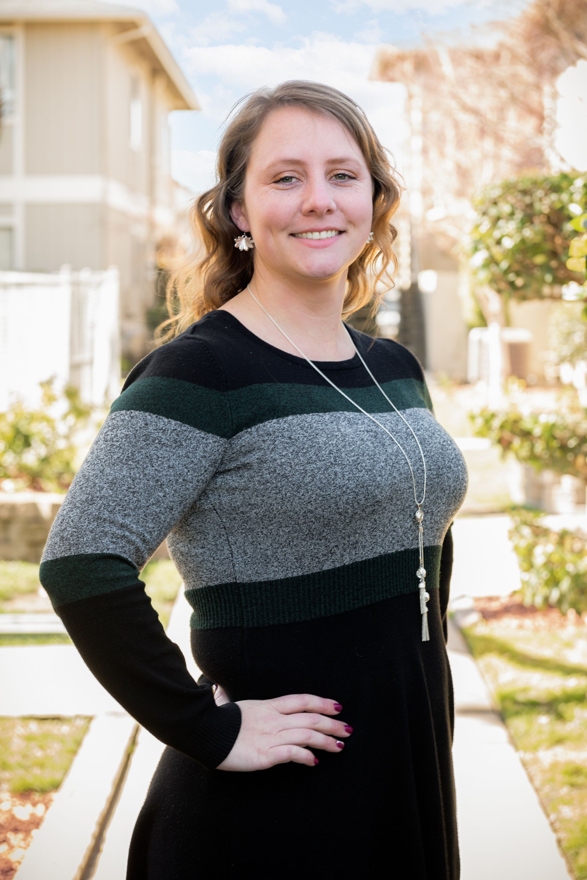 Woman with hand on hip smiling in front of apartment building, wearing sweater with green and gray stripes.