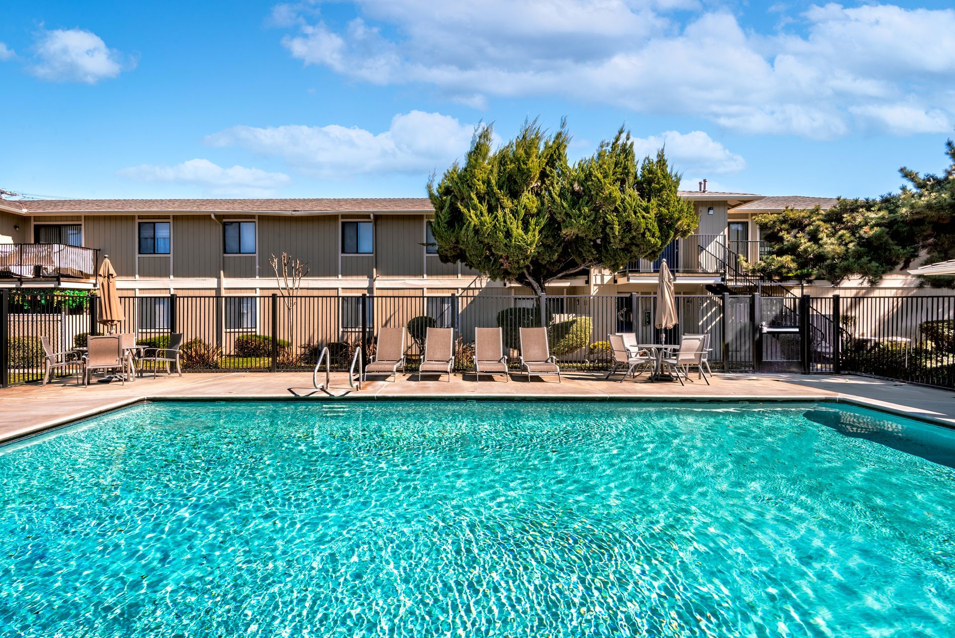 Swimming pool in front of a two-story building. Lounge chairs and a tree are nearby under a blue sky.