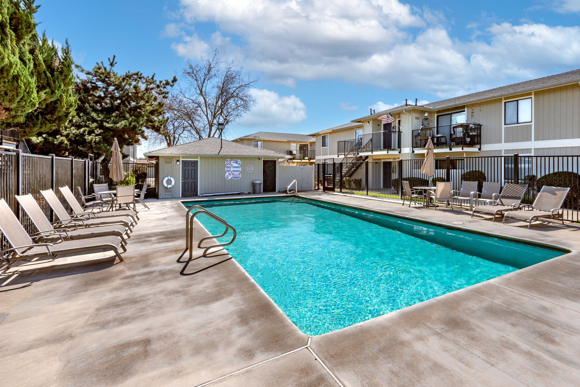 Pool with lounge chairs, surrounded by apartments on a sunny day.
