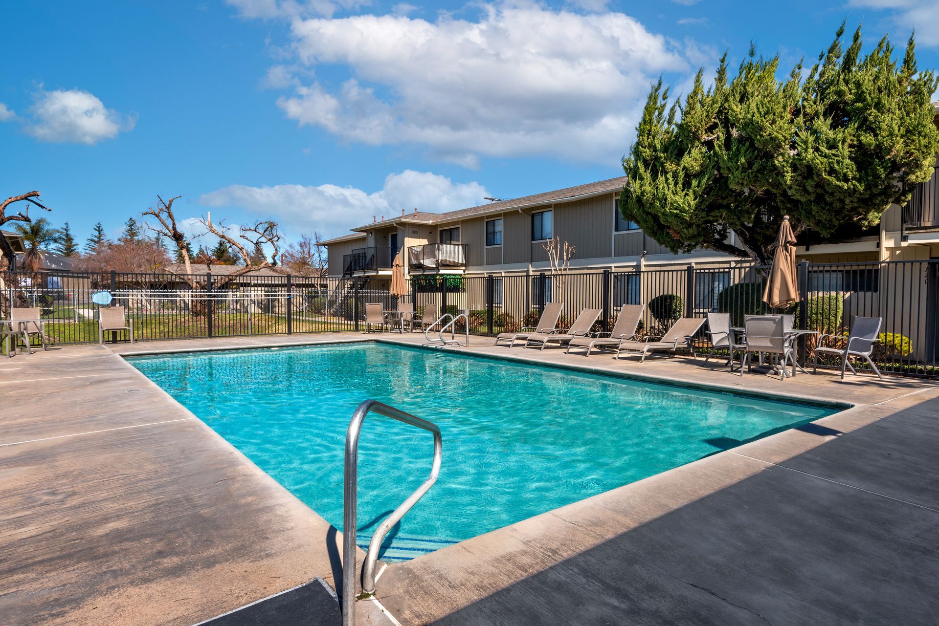 Poolside apartment complex on a sunny day with a blue pool, lounge chairs, and sky.
