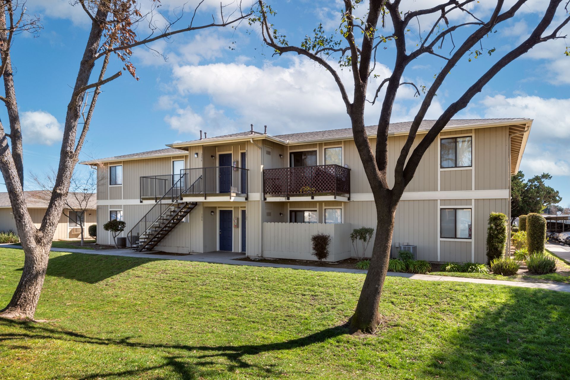 Two-story apartment building with light siding, green grass, and trees under a blue sky.