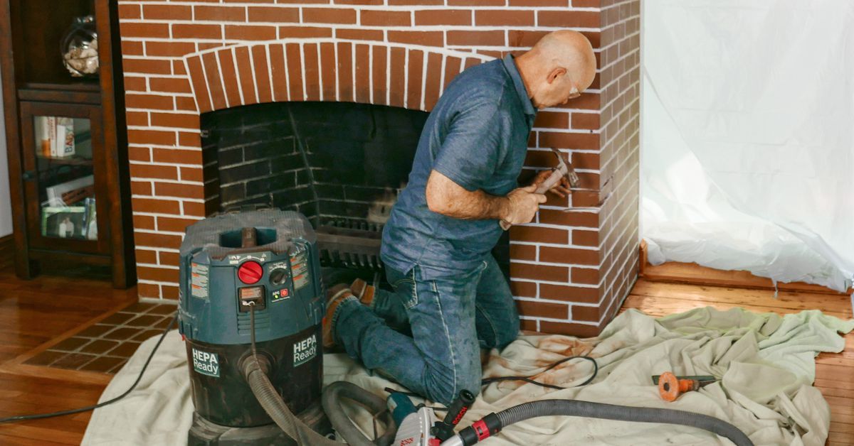 A man is kneeling in front of a fireplace with a vacuum cleaner.