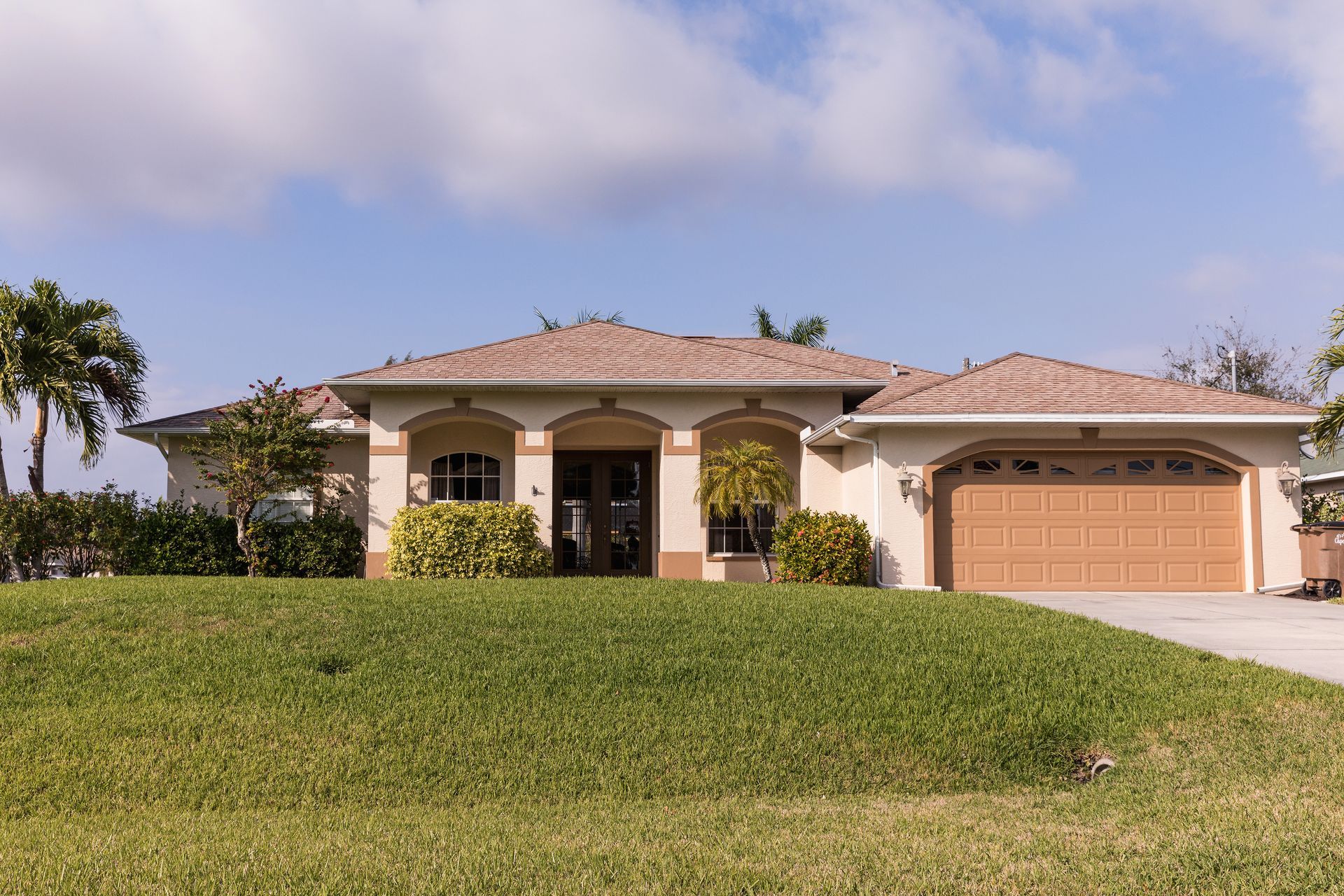 Single-story suburban house with a tiled roof, large lawn, and a two-car garage under a partly cloudy sky