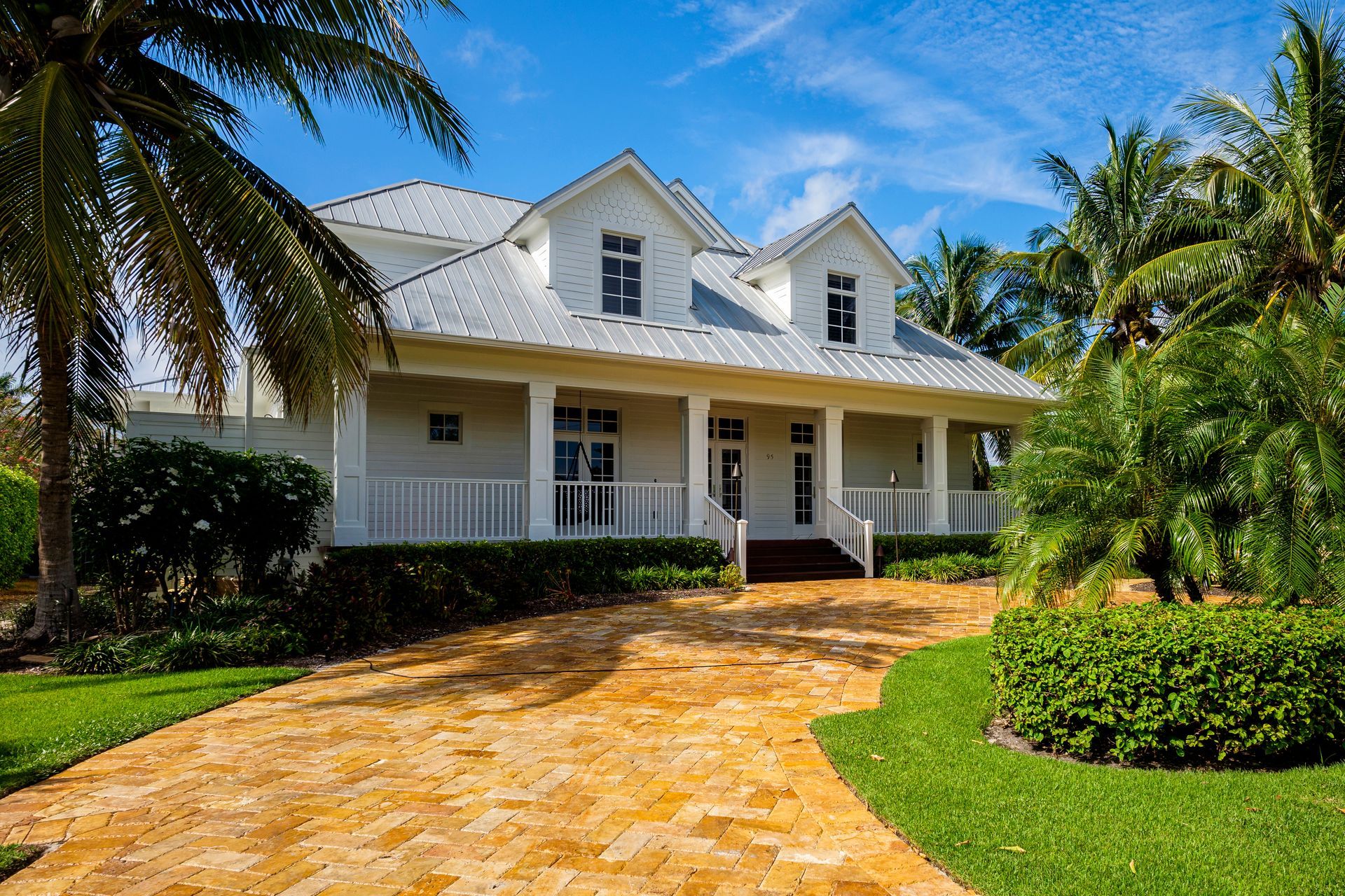 White house with a porch and palm trees, fronted by a curved stone driveway and lush green landscaping