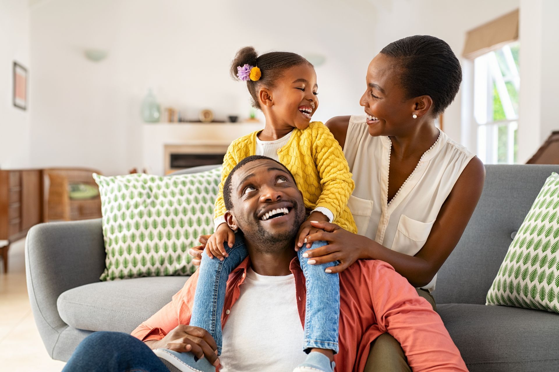 Family smiling together on a couch in a bright living room, child sitting on an adult’s shoulders