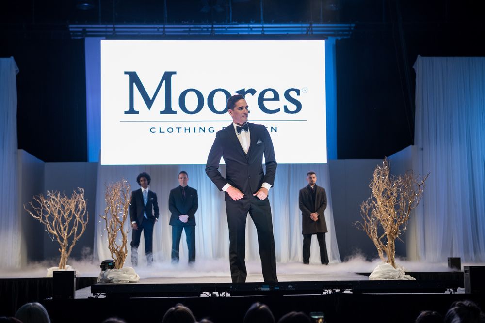 A man in a tuxedo is standing on a stage in front of a moores clothing sign.