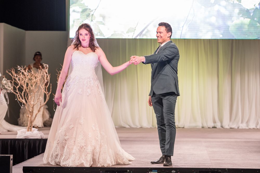A bride and groom are holding hands on a stage at a wedding show.