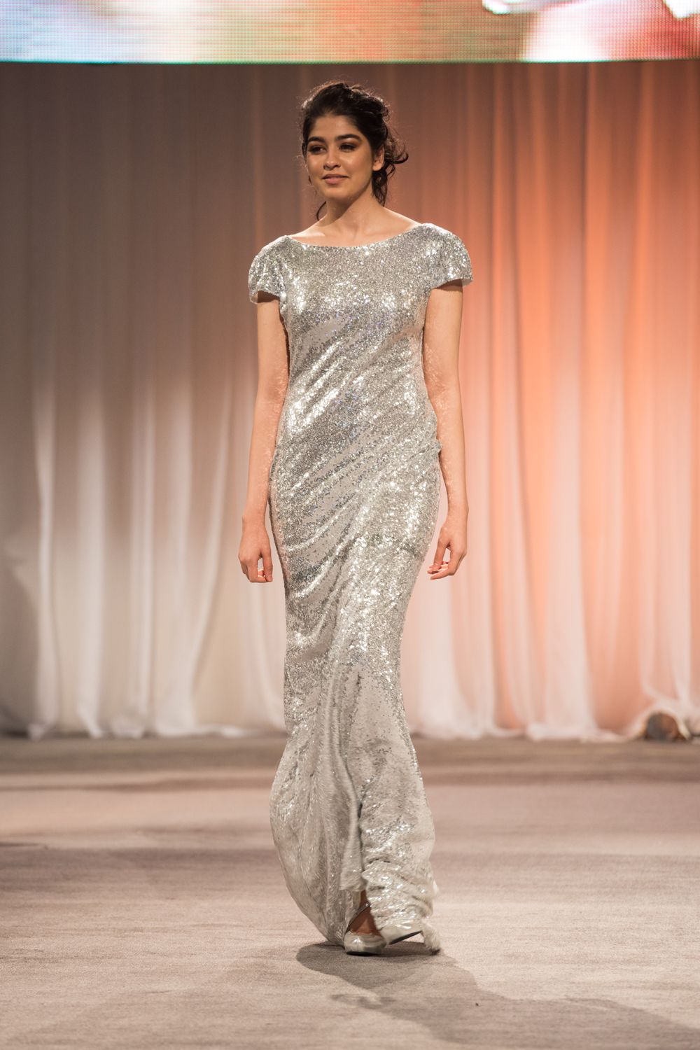 A woman is walking down a runway wearing a silver sequined dress.