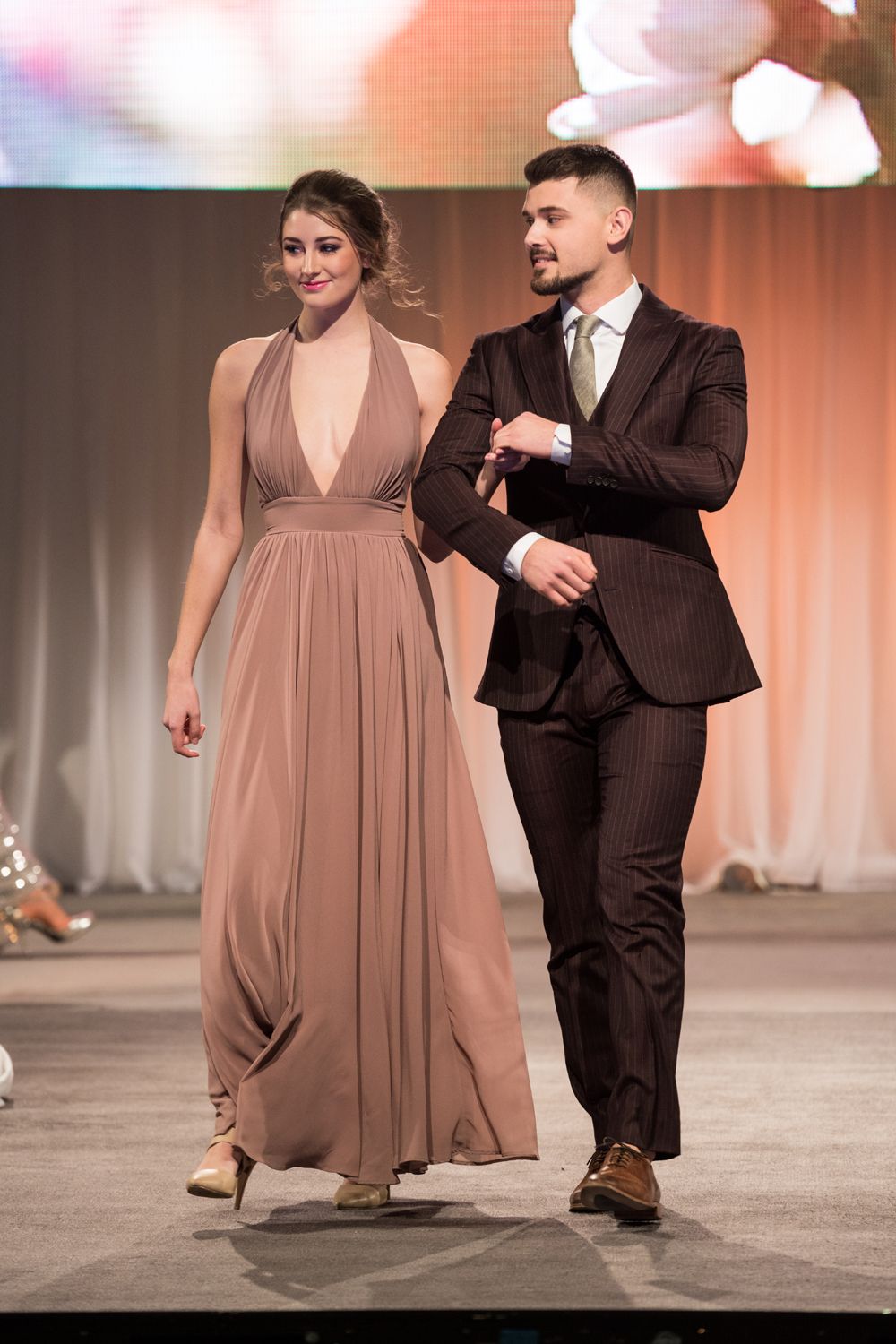 A man in a suit and a woman in a dress are walking down a runway.