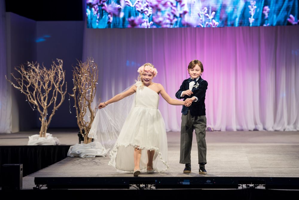 A boy and a girl are walking down a runway at a fashion show.