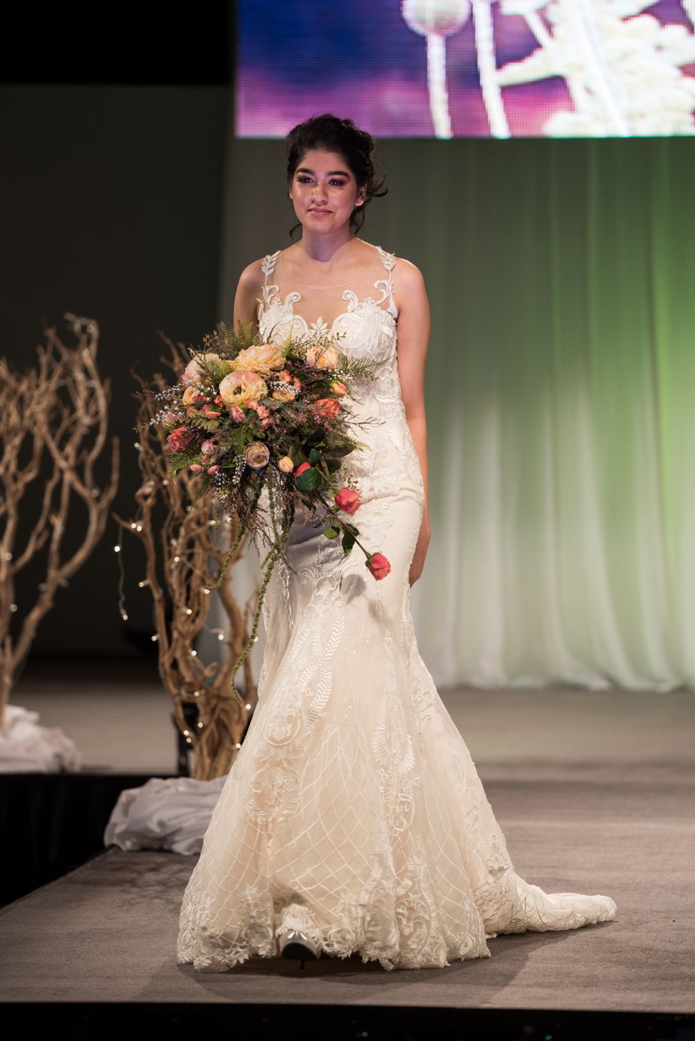 A woman in a wedding dress is walking down a runway holding a bouquet of flowers.