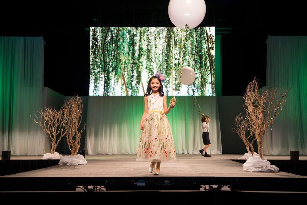 A little girl is walking down a runway holding a balloon.