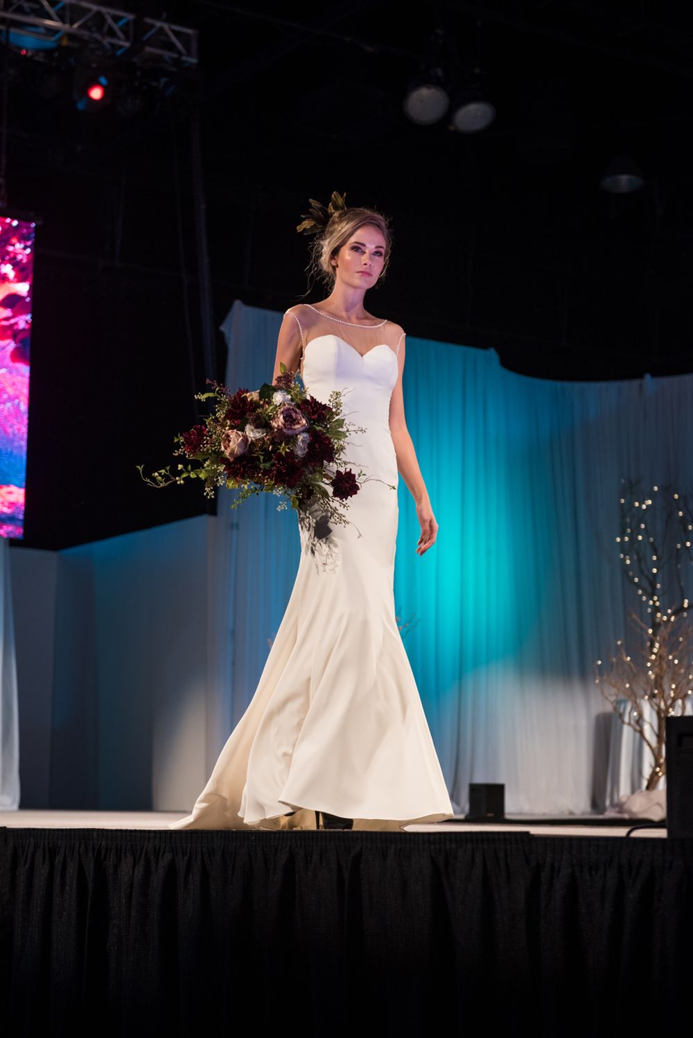 A woman in a wedding dress is walking down a runway holding a bouquet of flowers.