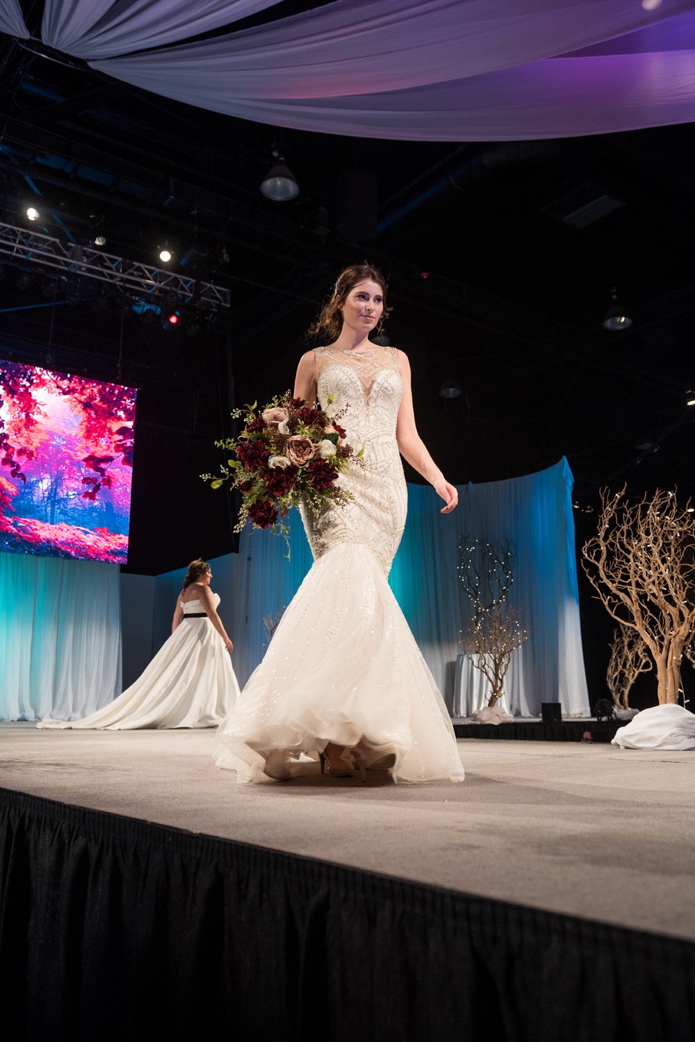 A woman in a wedding dress is walking down a runway holding a bouquet of flowers.