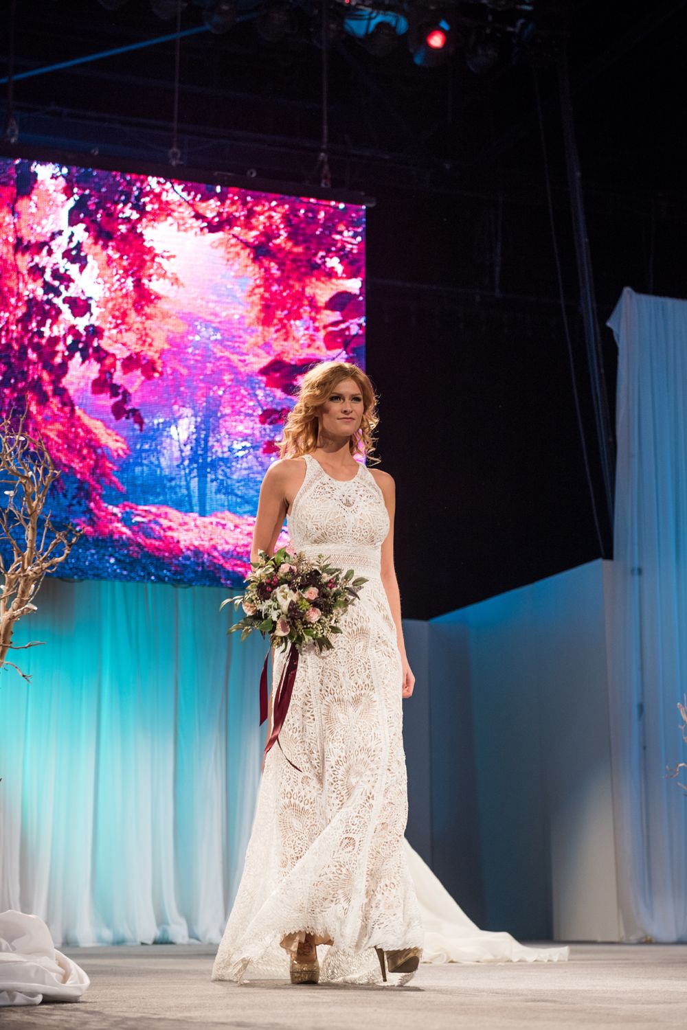 A woman in a wedding dress is walking down a runway holding a bouquet of flowers.