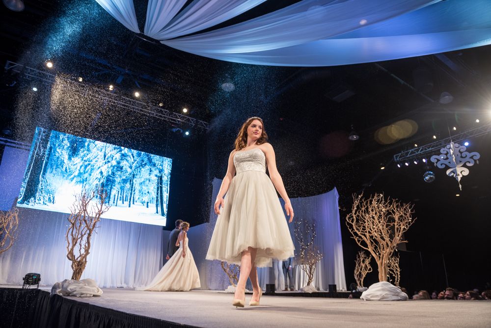 A woman in a white dress is walking down a runway at a wedding show.
