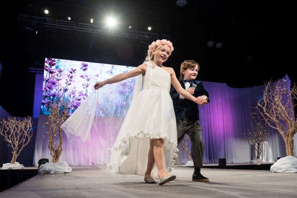 A boy and a girl are walking down a runway at a wedding show.