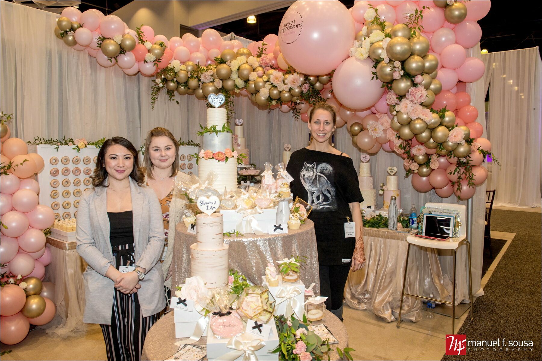 Three women are standing in front of a table with a cake and balloons.