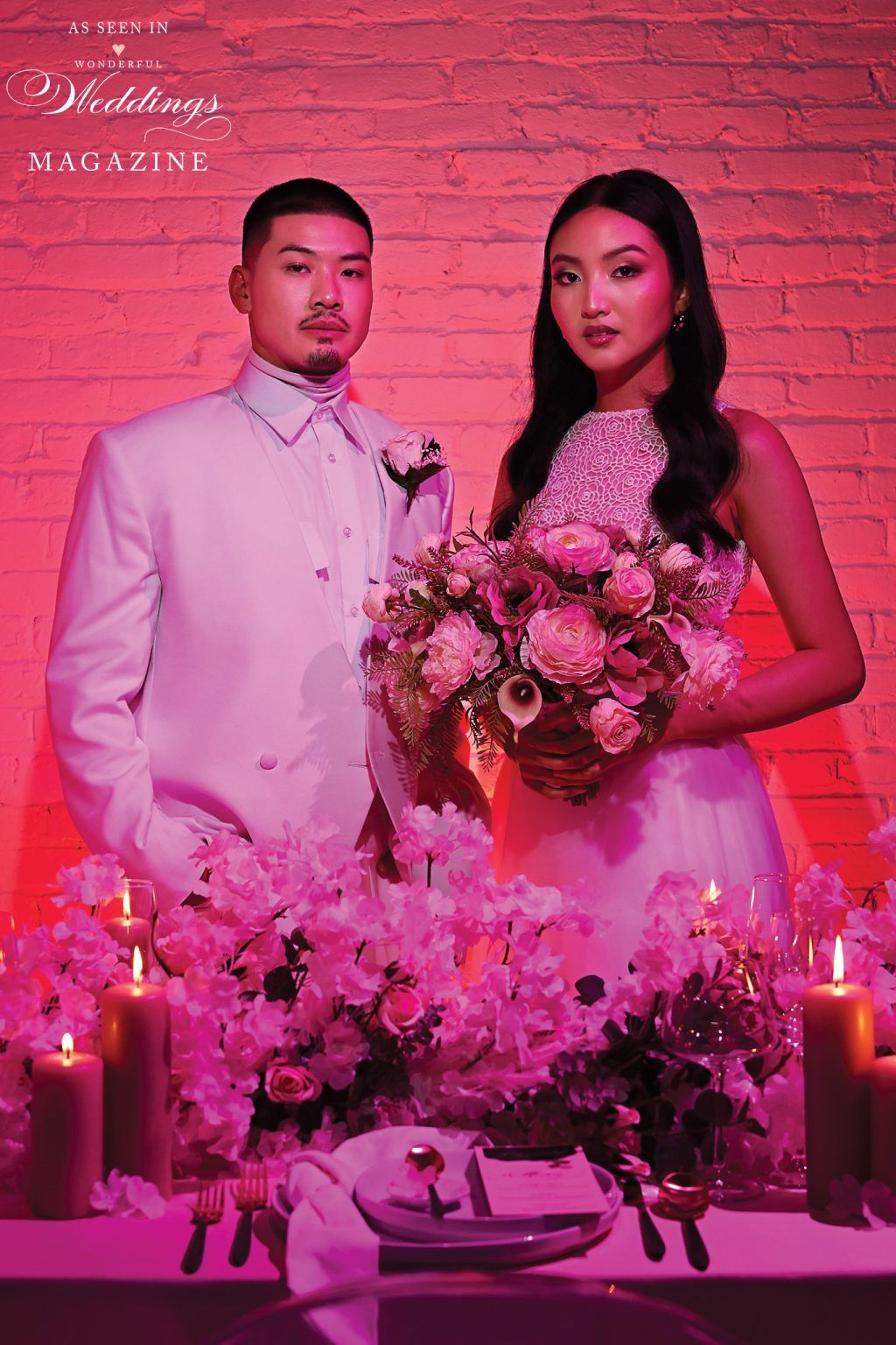 A bride and groom are posing for a picture in front of a table with pink flowers and candles.
