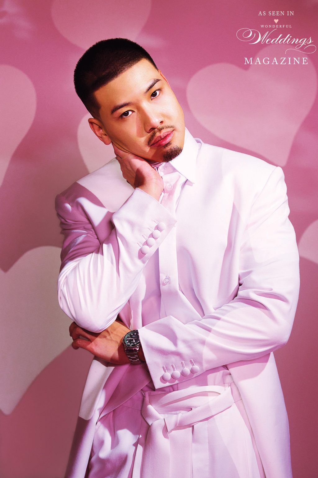 A man in a white suit is standing in front of a pink background with hearts.