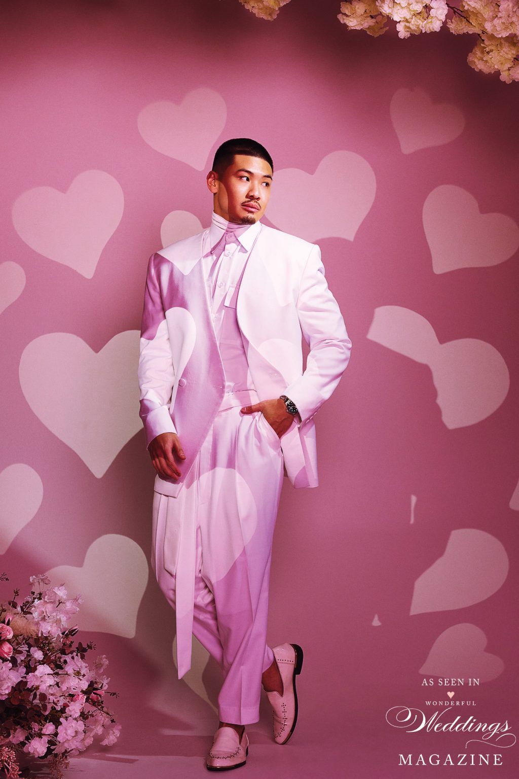 A man in a white suit is standing in front of a pink background with hearts.