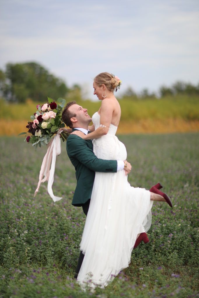 A bride and groom are standing in a field and the groom is holding the bride in his arms.