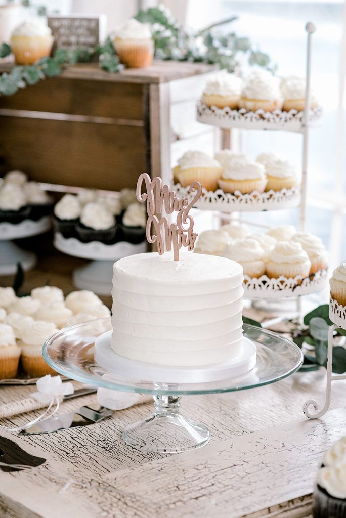 A white cake is sitting on top of a glass plate on a table surrounded by cupcakes.