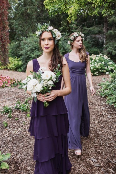 Two bridesmaids wearing purple dresses and flower crowns are walking in a garden.